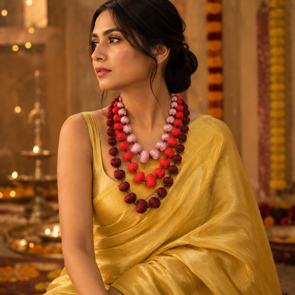 A woman wearing a yellow saree and a layered beaded modifiable Aditi necklace with red, pink, and maroon beads, posed in festive setting.