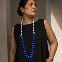 A woman wearing a long multi-strand handmade fabric beaded necklace with light blue, turquoise, and royal blue beads, paired with a black sleeveless top.