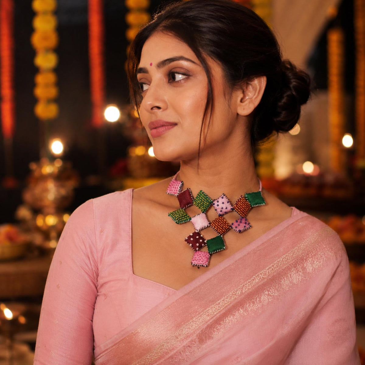 A woman wearing a pink traditional outfit and a colorful Adwait neckpiece which is made of several square pillows, photographed in a warmly lit festive indoor setting.