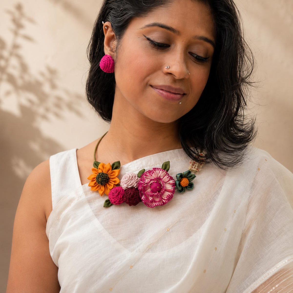 A woman wearing a white saree and a colorful floral statement Gul necklace with pink, orange, and green embroidered flowers on a metal band.
