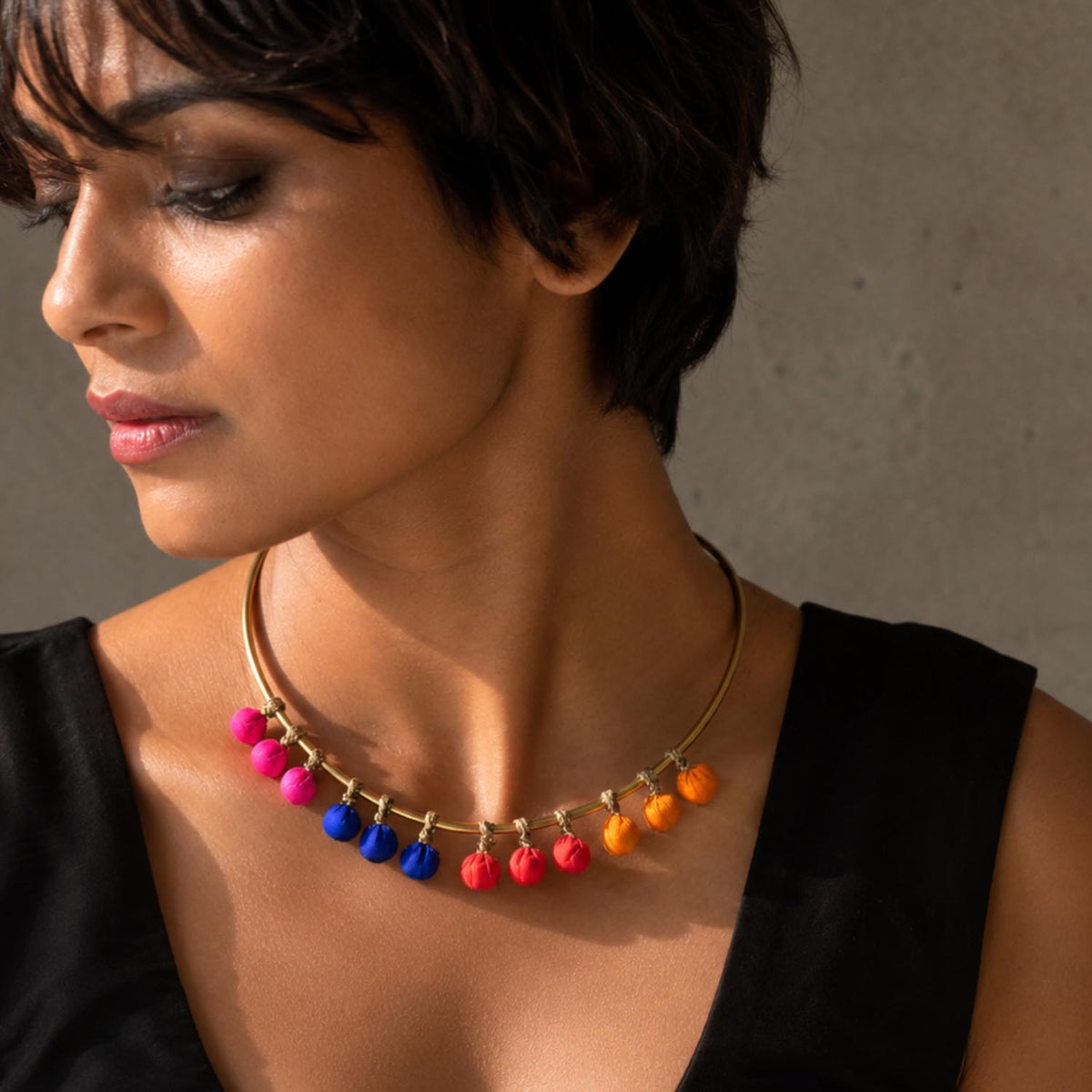 A woman wearing a brass necklace with dangling colorful beads in pink, blue, red, and yellow paired with a black sleeveless top against a neutral gray background.