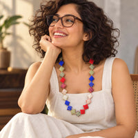 A woman with curly brown hair and glasses smiling while wearing a colorful handmade Keya neckpiece made with square textile pillows. 