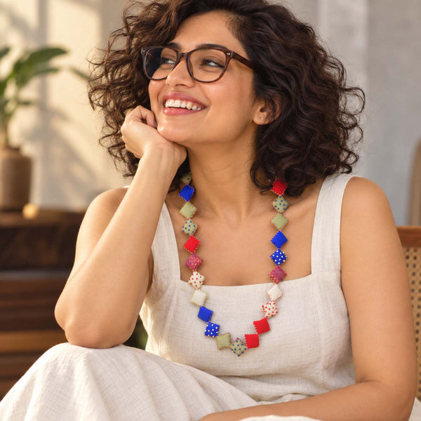 A woman with curly brown hair and glasses smiling while wearing a colorful handmade Keya neckpiece made with square textile pillows. 