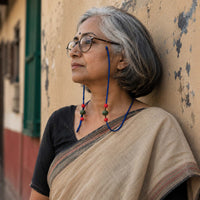 A woman with gray hair wearing specs attached with Khushi Specs retainer with red and green bead beads, leaning against a textured beige wall.