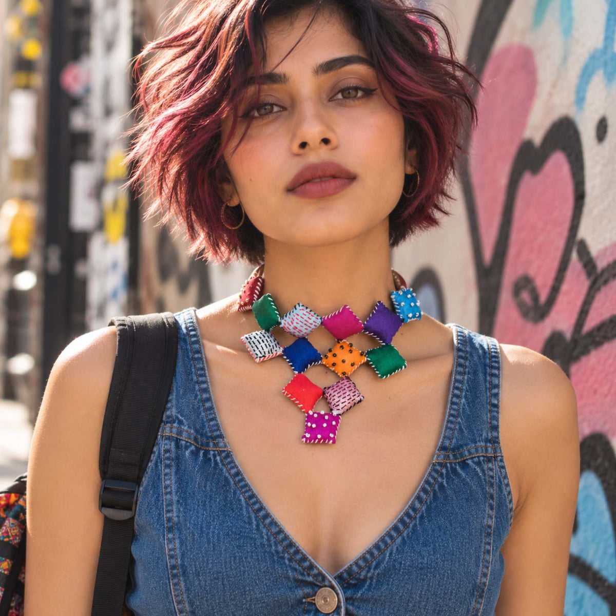 A woman wearing the Adwait colorful geometric textile necklace over a denim top, standing in front of a graffiti-covered wall.