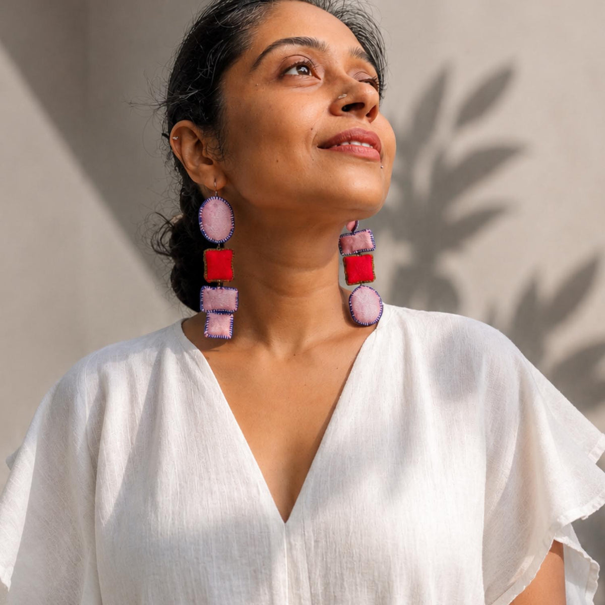 A woman wearing Nirali dangle earrings in pink, red, photographed in warm natural sunlight against a neutral wall.