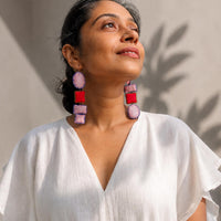 A woman wearing Nirali dangle earrings in pink, red, photographed in warm natural sunlight against a neutral wall.