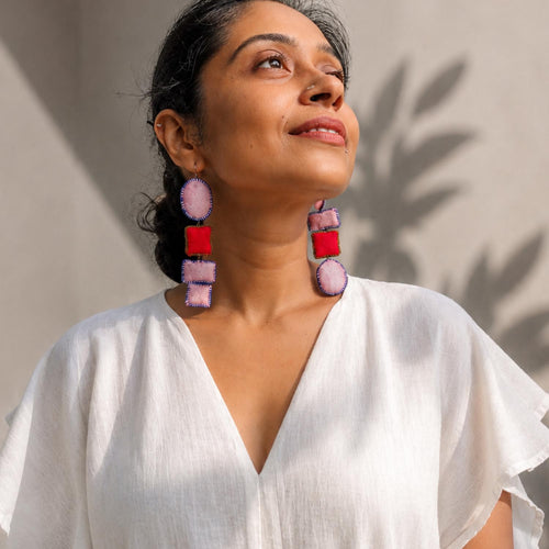 A woman wearing Nirali dangle earrings in pink, red, photographed in warm natural sunlight against a neutral wall.