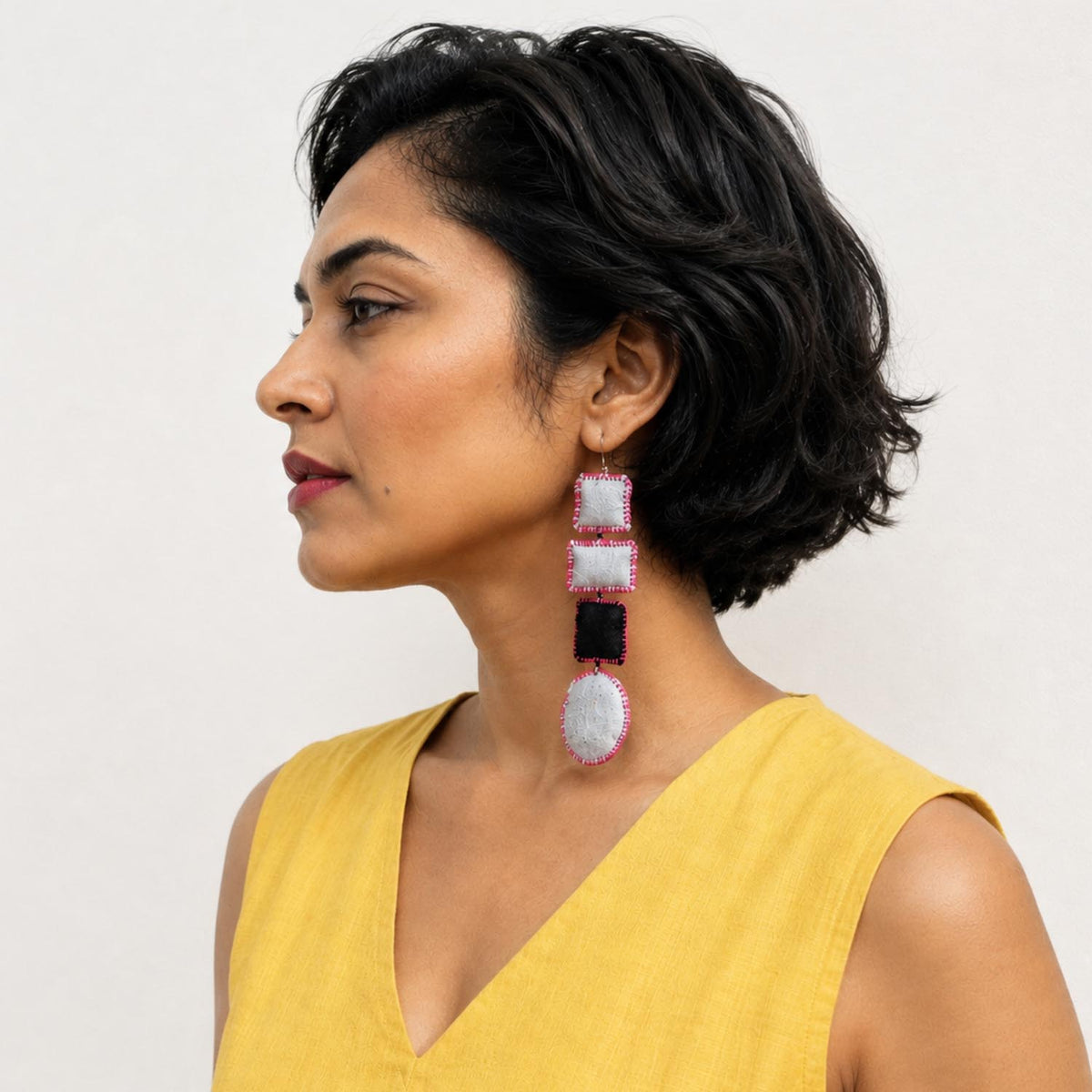 A woman in a yellow sleeveless top shown in profile, wearing an asymmetric Nirali earrings in grey and Black fabric pillow shapes.