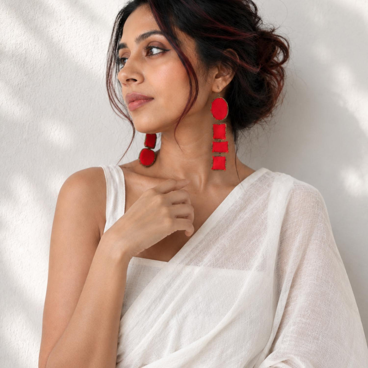 A woman wearing dramatic red geometric Nirali drop earrings with stacked circular and rectangular shapes, paired with a white saree against a softly lit white wall.