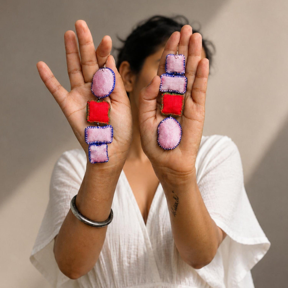 A person holds up both hands to display a pair of asymmetric Nirali handmade earrings. The earrings feature pink and red fabric-like rectangular and oval pieces , shown against a neutral beige background.