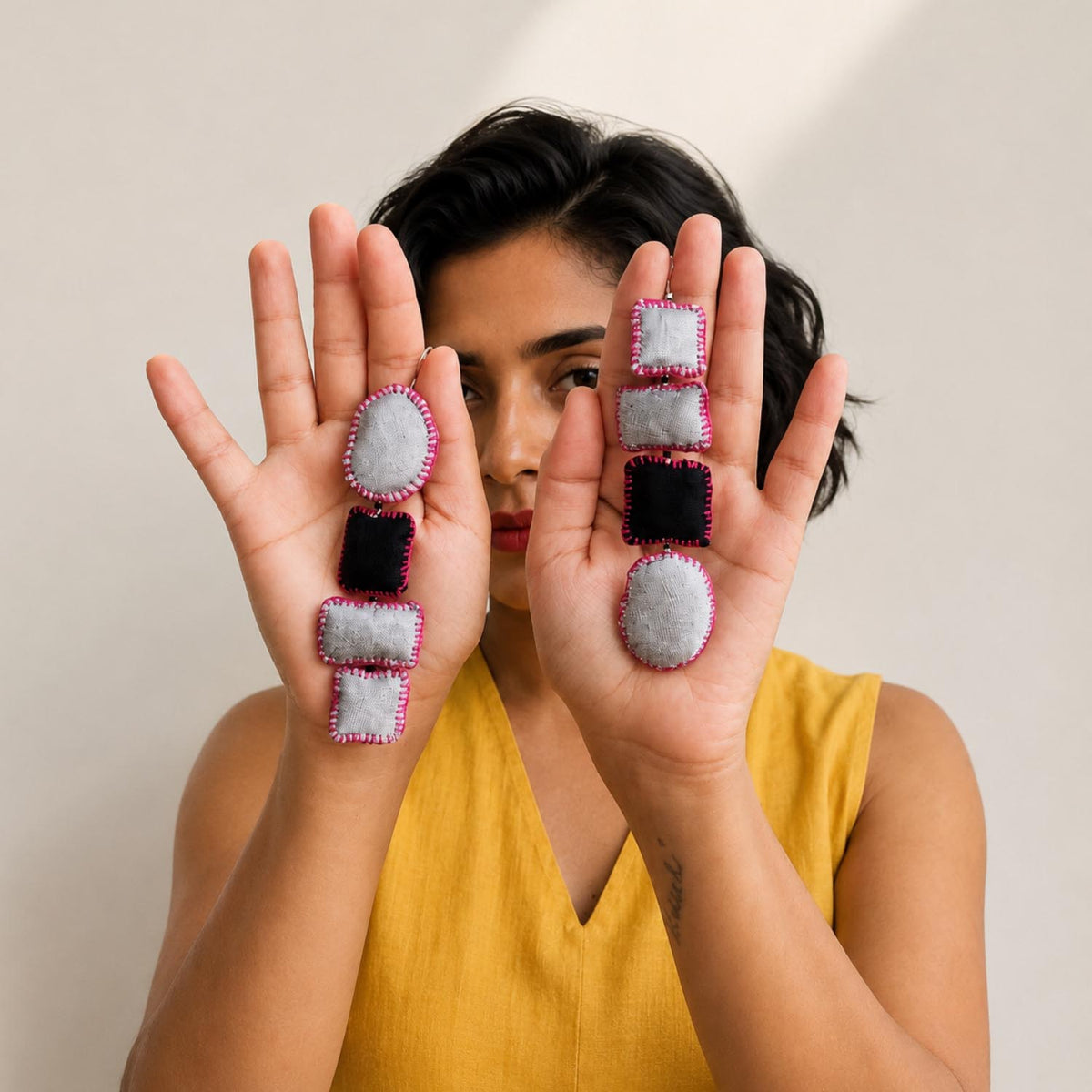 A woman in a yellow top holds up two hands displaying the statement fabric Nirali earrings which is handmade and uses different shapes of fabric pillows.