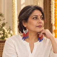 A woman wearing long beaded dangle earrings in red, white, and blue, posed in a warm, decorated indoor setting.