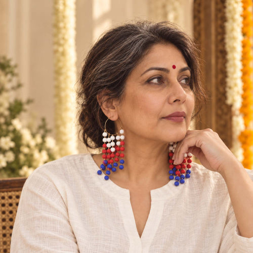 A woman wearing long beaded dangle earrings in red, white, and blue, posed in a warm, decorated indoor setting.