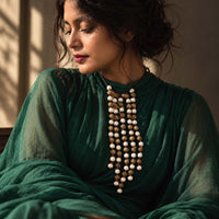 A woman wearing a dark green traditional outfit and a long multi-strand Noor beaded necklace with white and brown beads, captured in warm, directional sunlight.