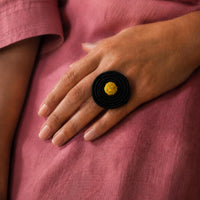 A close-up of a hand wearing a black spiral ring - handcrafted Rangoli ring with a small yellow center, set against a pink fabric background.