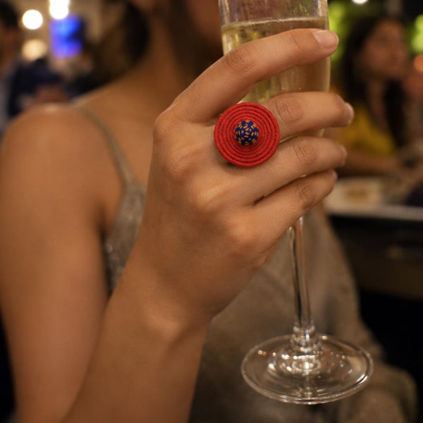 A close-up lifestyle shot of a person holding a glass of champagne, wearing Rangoli spiral fabric ring with a blue and yellow embroidered center on their finger.