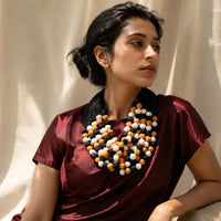 A woman wearing Ratna statement necklace with yellow, white, and black beads over a deep burgundy satin top, posed against a draped beige fabric backdrop.