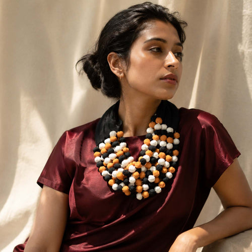 A woman wearing Ratna statement necklace with yellow, white, and black beads over a deep burgundy satin top, posed against a draped beige fabric backdrop.