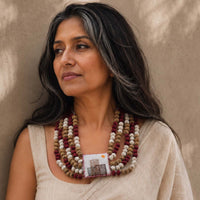 A woman with dark and gray hair wearing a multi-strand beaded Rekha necklace with a central fabric handembroidered pillow pendant paired with a beige saree against a textured beige wall.