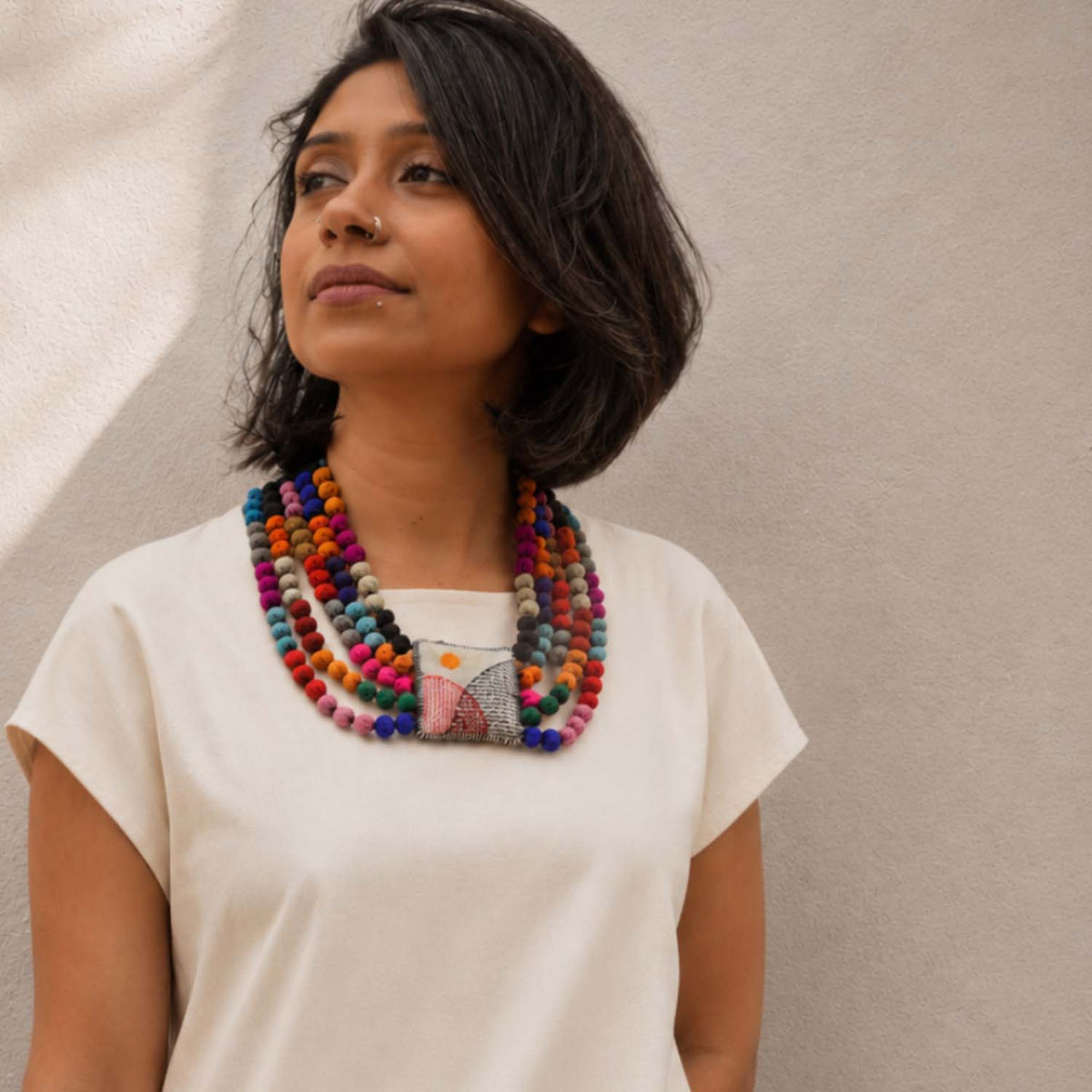 A woman with short dark hair wearing a cream-colored t-shirt and a thick, multi-colored Rekha Handmade necklace with a square handembroidered fabric pillowy pendant, standing against a textured beige wall.