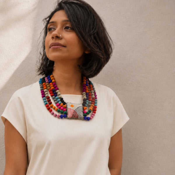 A woman with short dark hair wearing a cream-colored t-shirt and a thick, multi-colored Rekha Handmade necklace with a square handembroidered fabric pillowy pendant, standing against a textured beige wall.