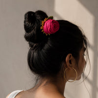 A woman shown from the back and side wearing a handcrafted hot pink fabric rosy hair accessory with small gold accents, styled in a bun, against a neutral wall with soft window shadows.