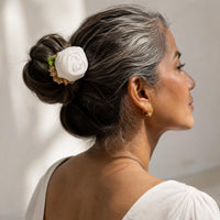 A woman shown in profile with her hair styled in a low bun, wearing a white handcrafted fabric flower Rosy bun stick hair accessory with a gold chain and green accent. She is wearing a white top against a softly lit, neutral background.