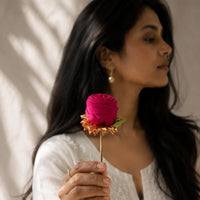 A woman with long dark hair holds a handcrafted vibrant magenta-pink fabric rose bun stick with a metal stem against a warm beige background with soft leaf shadows.