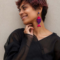 A woman with short burgundy hair wearing a black sheer top and a colorful fish-shaped earring with blue, purple, and pink patterns, posing against a neutral beige wall.