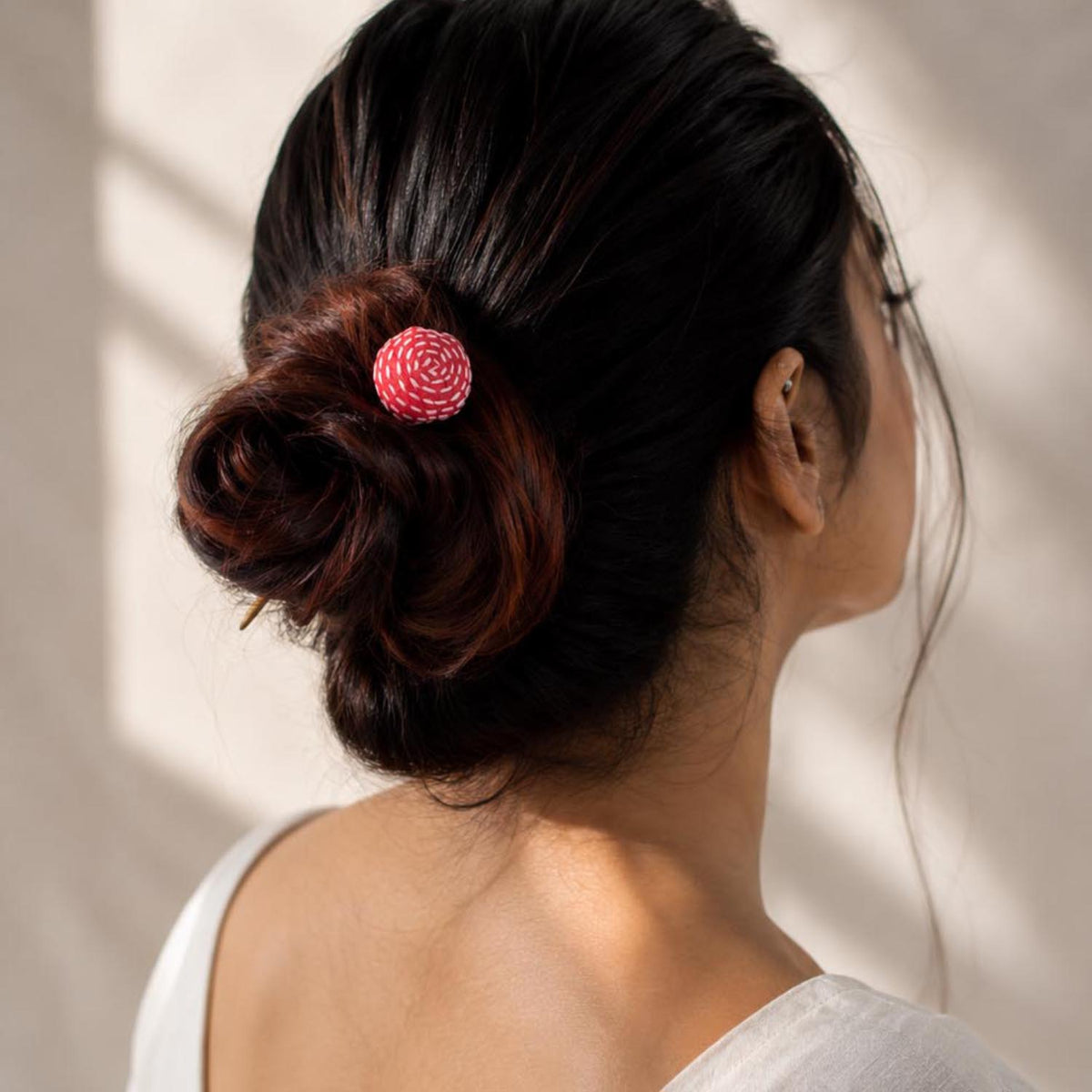 A woman shown from the back wearing a simple handcrafted Sarala fabric on metal bun stick hair accessory in her dark hair bun, set against a softly lit beige wall.