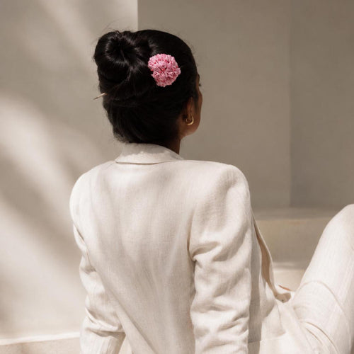 A woman seen from behind wearing a handmade Shiuli pink flower hair accessory in her dark hair, dressed in a white linen jacket and pants.