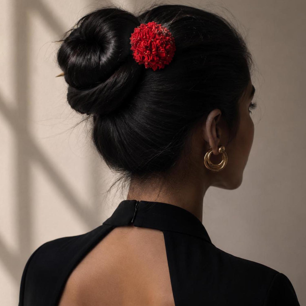 A woman shown from the back wearing a black dress with a high collar and open back, her dark hair styled in a bun with a bright red handcrafted textile Shiuli bun stick hair accessory. 