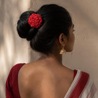 A woman shown from the back wearing a red floral handmade Shiuli hair accessory in her dark hair bun, with a red and white sari and a gold Jhumka earring, set against a warm beige wall with soft shadows.
