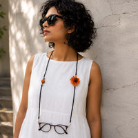 A woman wearing shades on her eyes and another sunflower specs retainer attached to eyeglasses around her neck, posing against a textured light gray wall.