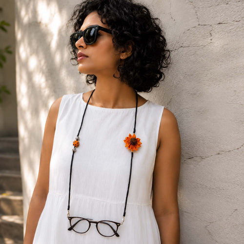 A woman wearing shades on her eyes and another sunflower specs retainer attached to eyeglasses around her neck, posing against a textured light gray wall.