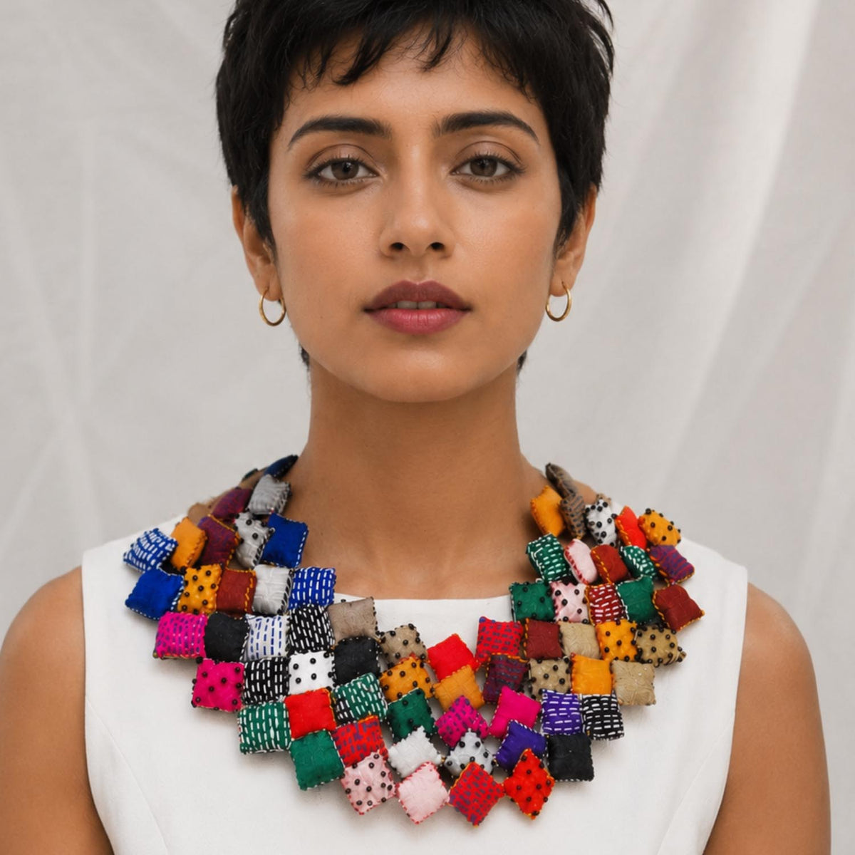 A woman with short dark hair wearing a large, Tejaswini  bib necklace made of many small fabric squares in different colors and patterns, paired with a white sleeveless top against a light draped background.