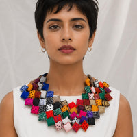 A woman with short dark hair wearing a large, Tejaswini  bib necklace made of many small fabric squares in different colors and patterns, paired with a white sleeveless top against a light draped background.