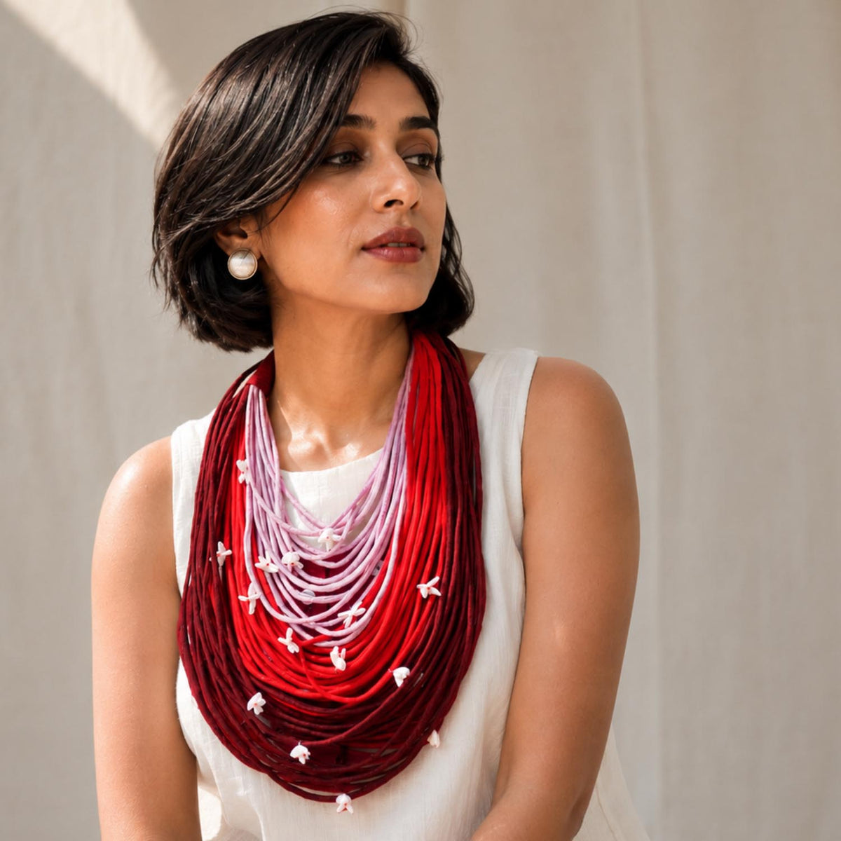 A woman wearing a multi-strand Vanya necklace with a red, pink, and maroon ombre gradient and small white floral shiuli accents, styled with a white sleeveless top against a neutral beige background.