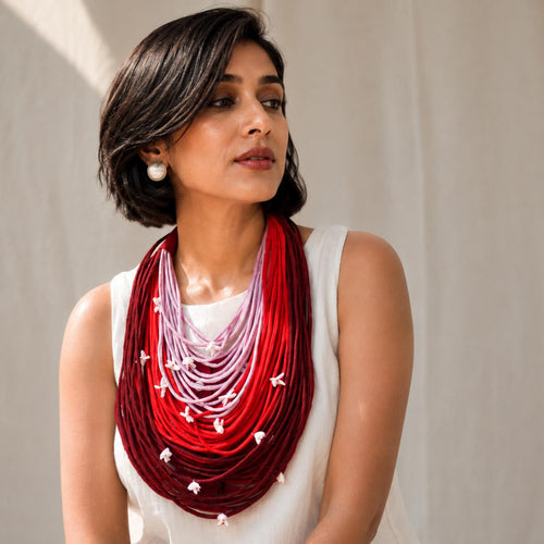 A woman wearing a multi-strand Vanya necklace with a red, pink, and maroon ombre gradient and small white floral shiuli accents, styled with a white sleeveless top against a neutral beige background.