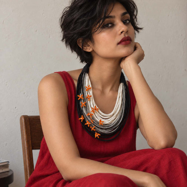 A woman with short dark hair wearing a handmade Vanya necklace in white, black, and gray fabric cords accented with movable small yellow fabric flowers.