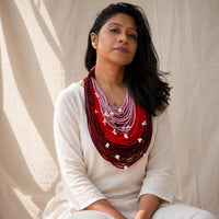 A woman wearing a statement Vanya -grand handcrafted necklace with a red, pink, and maroon ombre gradient and small white flower accents which are movable, styled with a cream-colored top against a draped beige fabric backdrop.