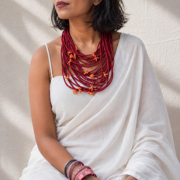 A woman wearing a Maroon Vanya Medium necklace with tiny adjustable yellow florals, styled over a white draped top against a textured off-white wall.
