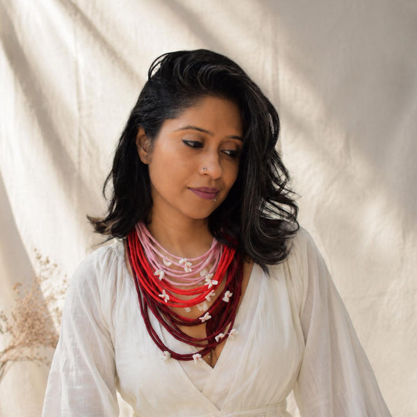 A woman with dark hair wearing a layered Vanya Medium handcrafted necklace set consisting of maroon, red and light pink cords with small white flower adjustable, movable charms  paired with a white blouse against a draped cream fabric backdrop.
