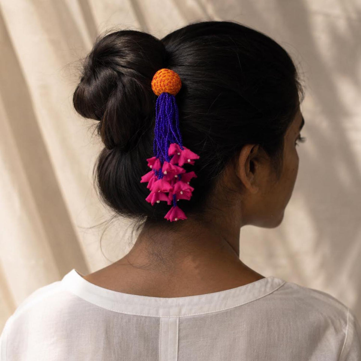 A woman shown from the back wearing a white top, with her dark hair styled in a low bun and secured with a handmade Zubeida hair accessory featuring an yellow round fabric bead, blue tassels, and bright pink flower-like  accents.