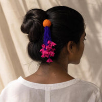 A woman shown from the back wearing a white top, with her dark hair styled in a low bun and secured with a handmade Zubeida hair accessory featuring an yellow round fabric bead, blue tassels, and bright pink flower-like  accents.
