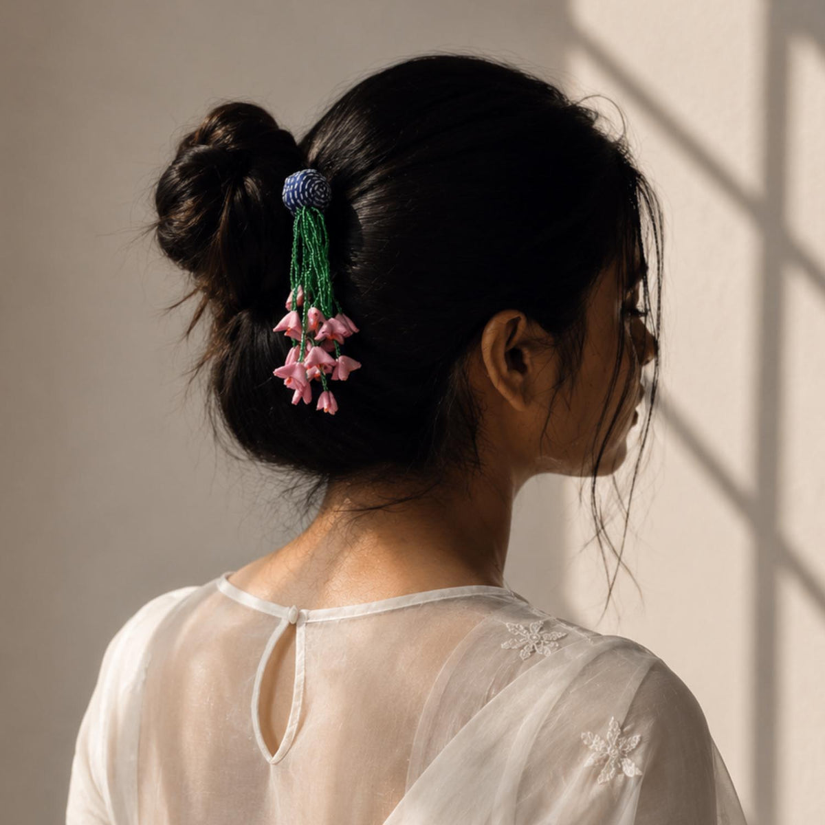A woman shown from the back wearing a dark bun secured with a Dangling handcrafted fabric zubeida bunstick featuring pink flower-like tassels and green strands, against a softly lit neutral wall.
