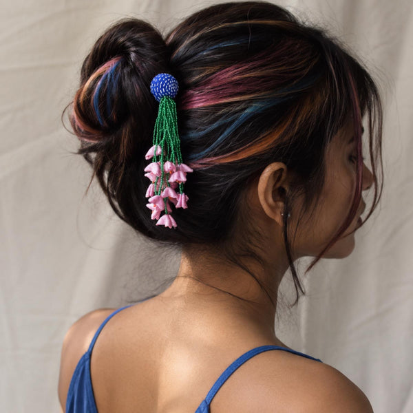 A woman shown from the back wearing a blue Zubeida fabric handcrafted bunstick hair accessory with dangling pink flower charms, styled in a bun against a light fabric backdrop.