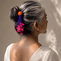 A woman with silver-gray hair styled in a low bun, wearing a colorful handmade fabric Zubeida Juda pin with a blue tassel, orange fabric bead top, and pink flower details. 