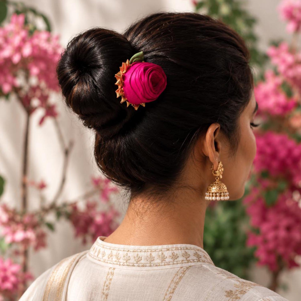 A woman shown from the back wearing a dark brown hair bun adorned with a bright pink hancrafted fabric rose juda pin hair accessory and a gold jhumka earring, set against a soft-focus background of pink flowers.